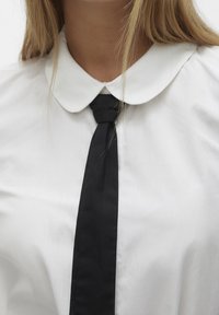 Chemise blanche à col avec une cravate noire. Texture de tissu lisse, design de col rond, et accent de cravate verticale centré sous le col.