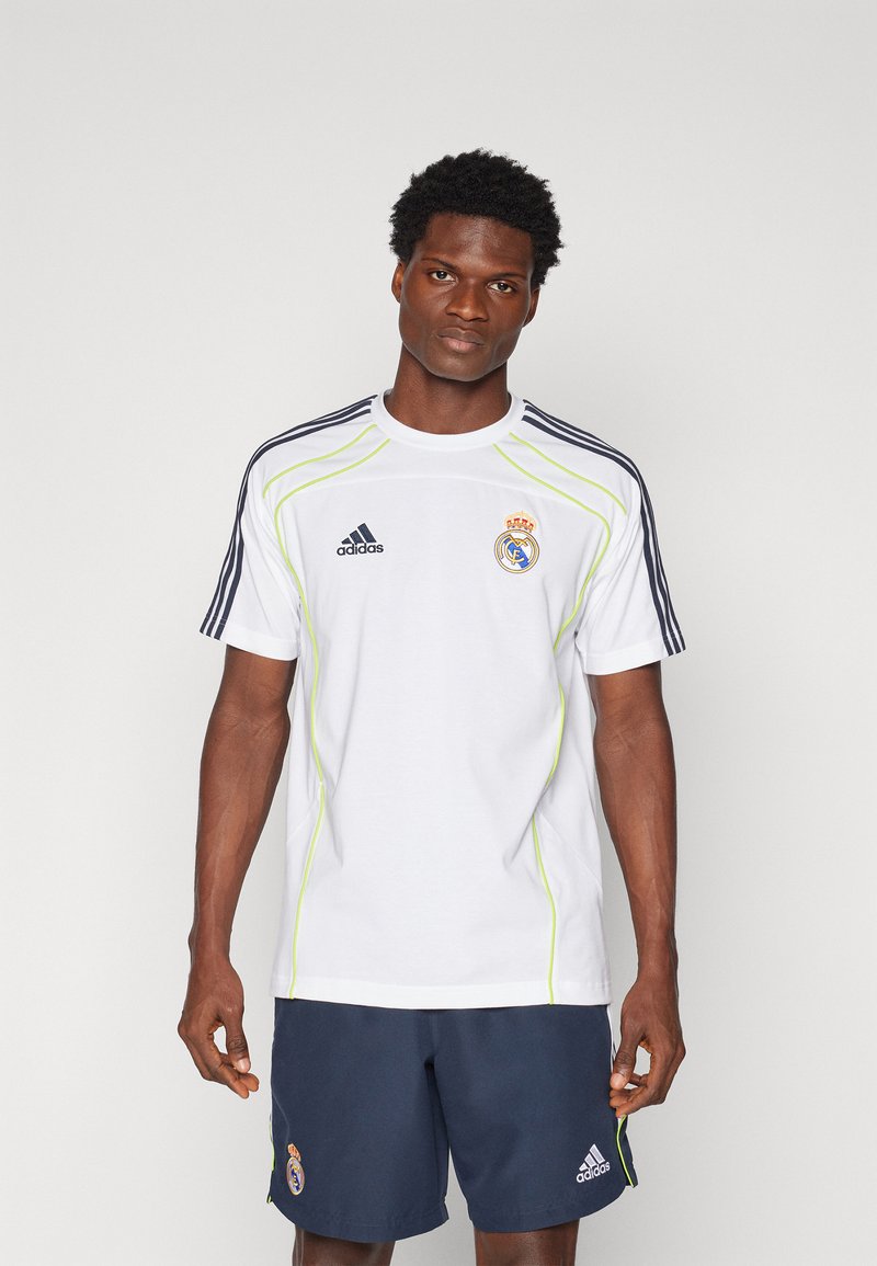 Camiseta atlética blanca con rayas negras, acentos verdes y logo de Real Madrid bordado. Usada con pantalones cortos oscuros que presentan un diseño similar.