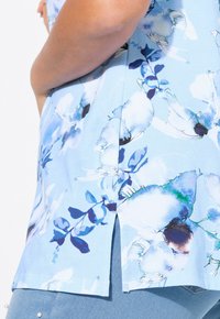 Blusa floral en azul claro con un diseño de acuarela que incluye acentos en blanco y azul oscuro, mangas cortas y aberturas laterales para mayor comodidad.