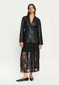 Zwarte leren blazer met contrasterende revers en een riem. Daaronder een doorschijnende zwarte kanten rok met bloemmotieven. Zwarte puntige ballerina's.