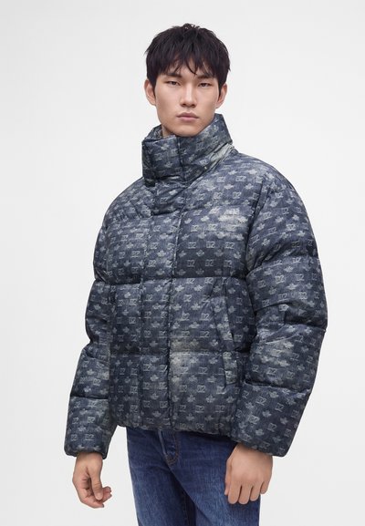 Jeune homme portant une doudoune bleue à motifs avec un col haut et un jean bleu, debout devant un fond blanc uni.