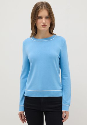 Vrouw draagt een lichtblauwe trui met lange mouwen en witte bies bij de kraag, mouwen en zoom, gecombineerd met een zwarte broek, staand tegen een witte achtergrond.