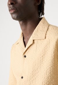 Short-sleeve, collared shirt in textured light beige fabric. Features a subtle pattern and dark buttons on the front.