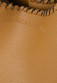 Žltá textúrovaná kožená taška s embosovaným logom "LOLACASADEI" a kontrastným čiernym prešívaním pozdĺž horného okraja.
