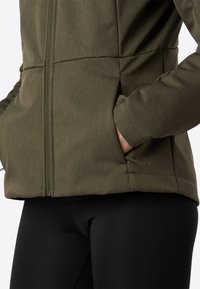 Olive green zip-up jacket with textured fabric, side pockets, and adjustable hem. Black leggings visible beneath.
