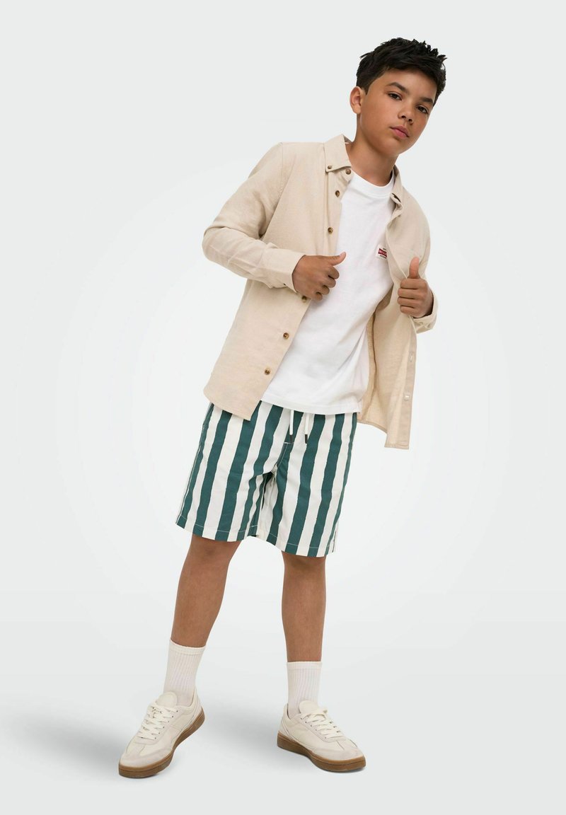 Garçon portant une chemise beige boutonnée sur un t-shirt blanc, un short à rayures vertes et blanches, des chaussettes blanches et des baskets beiges, debout avec les pouces levés.