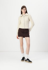 Sweater de malha creme com fecho zip na frente, combinado com uma mini saia azul-escura riscada a ferros e ténis pretos com detalhes brancos.