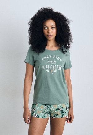 Grünes Baumwoll-T-Shirt mit verblasstem weißen Text "Très bien mon amour." Kombiniert mit kurzen Hosen mit blumigem Muster in grün, blau und orange.