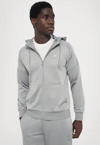 Grå zip-up hoodie lavet af blødt stof med en forlomma, snoretræk i hætten og logo-patch på brystet. En del af et matchende træningssæt.