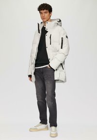 Witte gewatteerde jas met capuchon, zwarte ritsen en zijzakken, gedragen over een zwarte trui en grijze jeans; heeft een relaxte pasvorm.