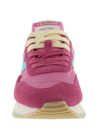 Vista frontal de un zapato deportivo rosa con cordones beige y rosa, detalle lateral azul claro y etiqueta "Victoria Since 1915" en la lengüeta.