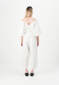 Blusa bianca off-the-shoulder con maniche a palloncino e laccio nella parte posteriore, abbinata a pantaloni bianchi a gamba larga, con scarpe nere.