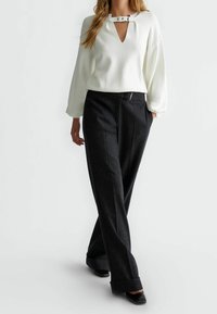 Femme portant un haut à manches longues blanc avec un décolleté carré et un pantalon large sombre, se tenant avec une main dans la poche.