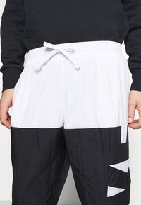 Mann in schwarzer und weißer Windbreaker-Hose mit an der Taille gebundenem Kordelzug, Hände an den Seiten, und schwarzem Langarmoberteil.