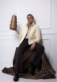 Kremowa polarowa kurtka zapinana na zamek błyskawiczny, z dwiema kieszeniami, noszona na jasnym topie. Modelka trzyma ułożone jedna na drugiej brązowe cylindryczne pudełka, siedząc na brązowej tkaninie.