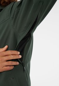 Groene softshell jas met een zijvak van mesh en een rits. De hand is boven het vak geplaatst, wat het functionele ontwerp benadrukt.
