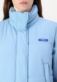 Šviesiai mėlynas paminkštintas liemenė, pagaminta iš minkštos medžiagos, su aukštu apykakle, priekiniais užtrauktukais ir mažuoju HUGO logotipu kairėje krūtinės pusėje.