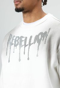 Witte trui met een "REBELLION"-logo in zilveren strasssteentjes met een druipend ontwerp. Ronde hals en gestructureerde stof.