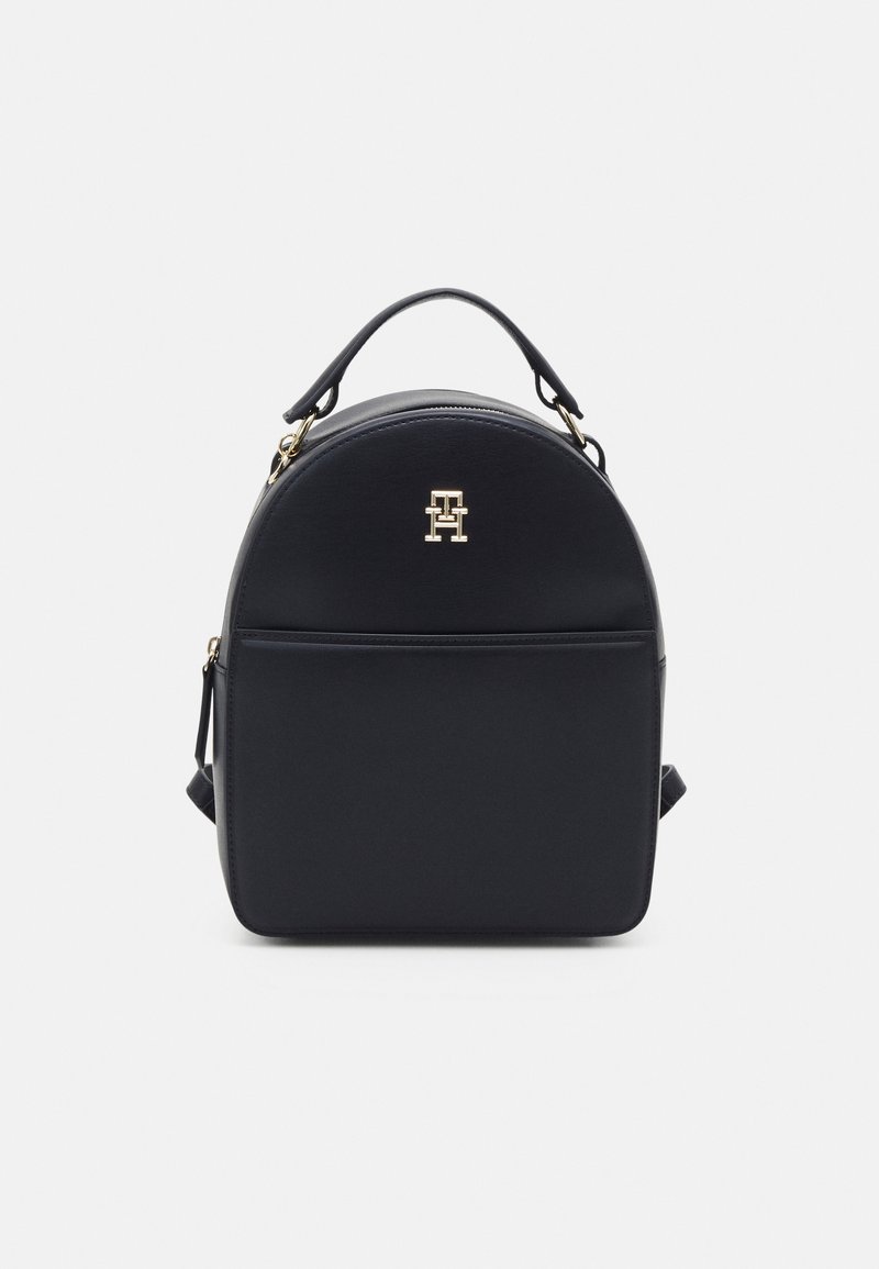 Sac à dos compact en cuir noir avec poignée supérieure, poche avant, ferrures dorées et petit logo métallique sur la partie avant supérieure centrale.