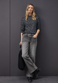 Pull en imprimé léopard noir et gris, jean gris à coupe décontractée et bottines noires. Le mannequin tient un sac noir.