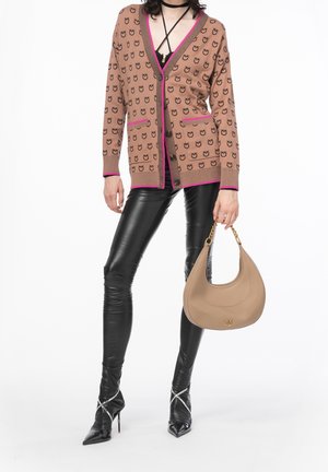 Femme portant un cardigan à motifs marron avec bordure rose, des leggings en cuir noir, des escarpins noirs pointus, tenant un sac à main beige avec une anse chaîne dorée.