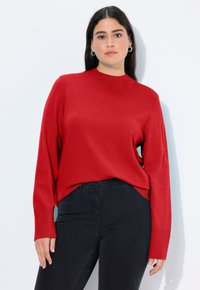 Maglione rosso lavorato a maglia con maniche lunghe e collo alto, con polsini a coste e una vestibilità morbida. Indossato con jeans neri.