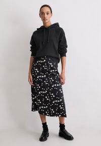 Felpa nera con cappuccio e lacci, abbinata a una gonna nera di lunghezza media con un motivo floreale bianco. Il modello indossa calze nere e scarpe.