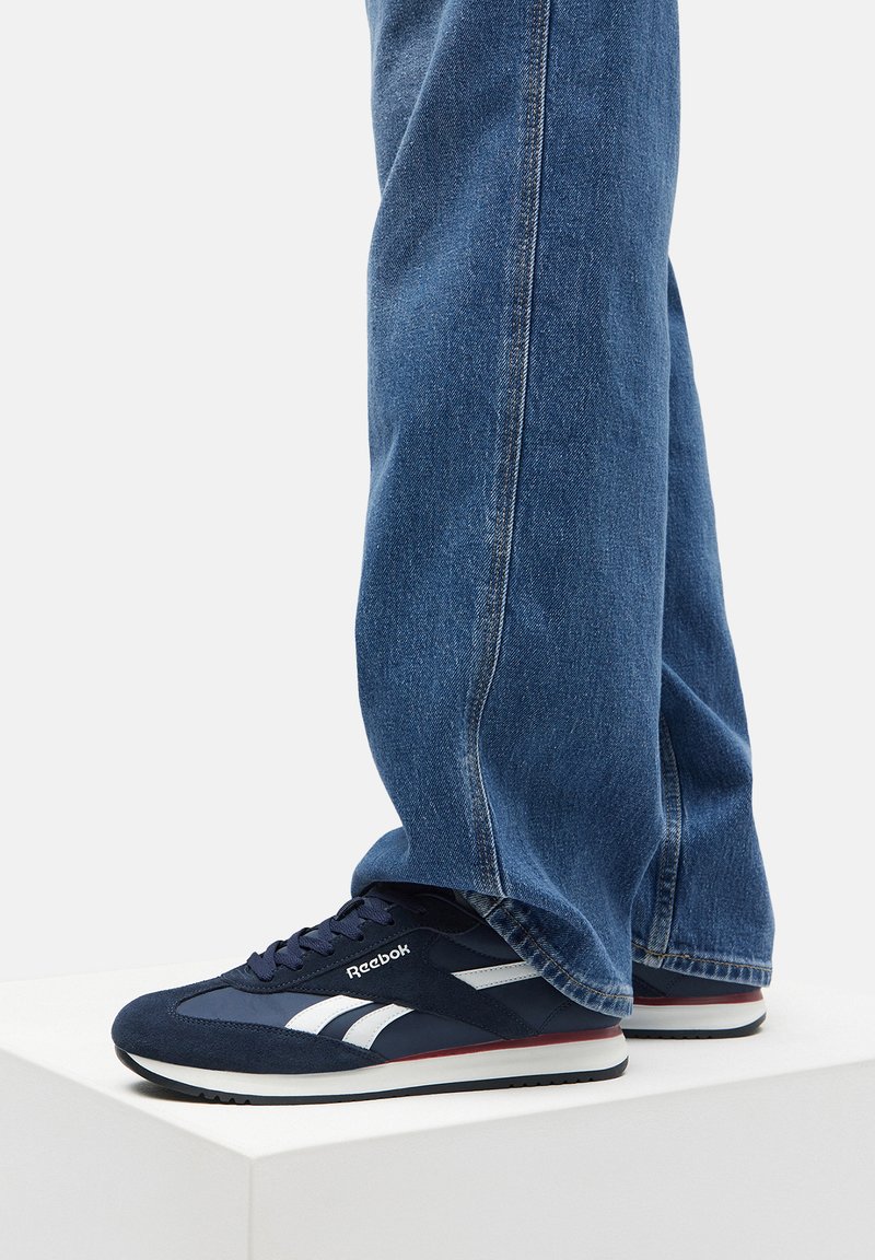 Granatowe sneakersy Reebok z zamszowej cholewki, białymi akcentami i białą podeszwą, zestawione z niebieskimi dżinsami.