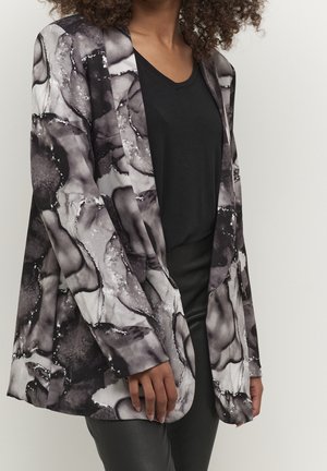 Kvinde iført sort og grå blazer med abstrakt mønster over en sort V-hals skjorte og sorte bukser, stående mod en ensfarvet baggrund.