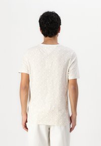 Kratkā piedurkne bežā T-kreklā ar ģeometrisku rakstu, izgatavots no mīksta auduma. Ierīkota ar apaļu kakla izgriezumu un taisnu apakšmalu.