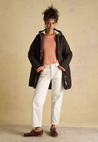 Parka imperméable marron foncé, doublure polaire blanche, pull à rayures orange, pantalon blanc taille haute, chaussures marron à lacets avec couture décorative.