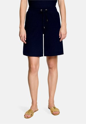 Femme portant un short noir mi-genou avec une taille à cordon et des sandales plates dorées, debout sur un fond blanc.