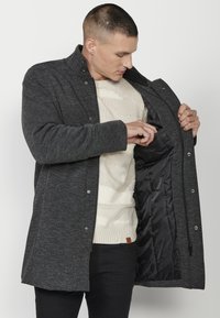 Abrigo gris texturizado con cuello alto y cierres a presión. Interior forrado con un bolsillo visible, sobre un suéter crema. Pantalones negros.
