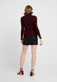 Pull à col roulé côtelé bordeaux à manches longues, associé à une mini-jupe en cuir noir. La personne porte des sandales à talons brunes.