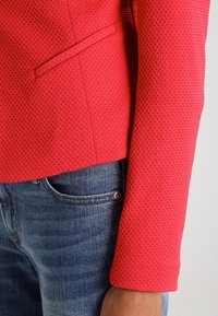 Rød tekstureret jakke med en tætsiddende design, der har en sidelomme, parret med blå denim jeans. Nærbillede af ærmet og stofmønster.