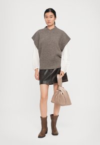 Frau trägt braunen Strickwesten über einer weißen Bluse, schwarzen Leder-Minirock, braunen Stiefeln und hält eine beige Knotenhandtasche vor einem einfarbigen Hintergrund.