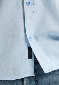 Světle modrá košile na knoflíky s jemnou texturou, s modrými knoflíky a černou etiketou na boku, v kombinaci s džíny.