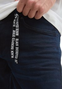 Tamne traperice s blagim teksturiranim završetkom. Sadrže crnu šnuru s natpisom "BLACK OVERDYED DENIM" u bijeloj boji.