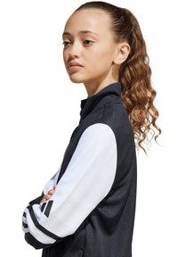 Schwarz-weiße Sportjacke mit hochgeschlossenem Kragen; verfügt über einen Reißverschluss vorne, Drei-Streifen-Akzente an den Ärmeln und strukturierte Stoffoberfläche.