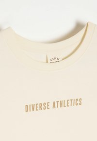 Kremowa bawełniana koszulka z rypsowym dekoltem w kształcie łódki. Na piersi znajduje się napis "DIVERSE ATHLETICS" w jasnobrązowym kolorze.