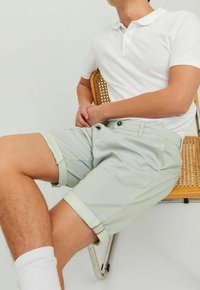 Světlé zelené kostkované šortky, ohrnuté manžety, měkká textura, nošeny s bílým polo tričkem. Osoba sedí na dřevěné židli.