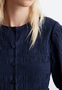 Navyblaues Kleid mit strukturierten Falten, kurzen Puffärmeln und einem runden Ausschnitt mit schwarzen Knöpfen. Glattes Gewebe.