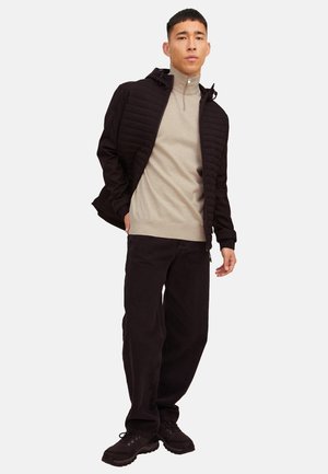 Mies, joka pukeutunut mustaan tikattuun takkiin beigen puoli-vetopaita-sweaterin, mustien housujen ja mustien lenkkareiden kanssa, seisoo valkoisella taustalla.