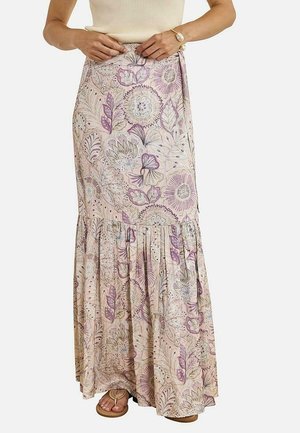 Mujer con falda larga beige floral con hojas moradas y verdes, combinada con sandalias y una blusa sin mangas color crema.