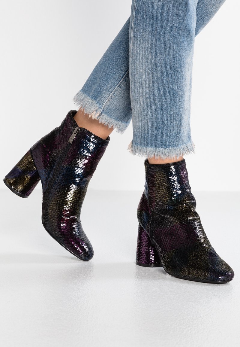 Botas negras hasta el tobillo con lentejuelas multicolores, acabado brillante y un tacón grueso. Cremallera lateral visible, combinadas con jeans de denim deshilachados.
