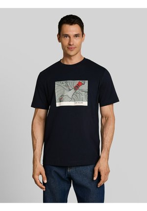 Mężczyzna noszący granatowy t-shirt z grafiką mapy topograficznej i czerwonym wzorem samochodu, w połączeniu z niebieskimi dżinsami, na jednolitym szarym tle.