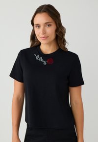 Vrouw die een zwart shirt met korte mouwen draagt, met een kleine rode roos en het woord "Valé" in het wit nabij de halslijn.
