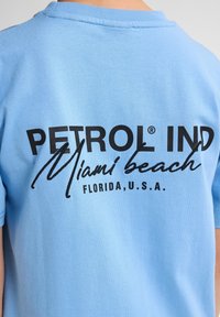 Světle modré bavlněné tričko s černě potištěným logem na zadní straně "PETROL IND Miami beach FLORIDA, U.S.A." s kontrastními texturami.