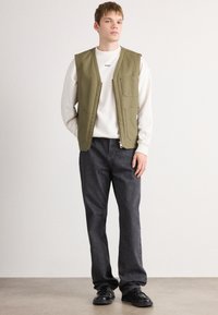 En ung mand står iført en olivengrøn vest over en hvid sweatshirt, løse sorte jeans og sorte lædersko foran en ensfarvet baggrund.