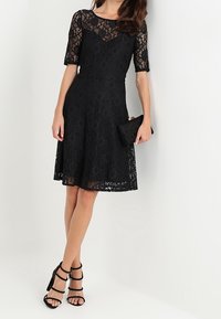 Robe en dentelle noire à manches courtes, jupe évasée et encolure ronde. Associée à des sandales à talons noires et à une petite pochette.